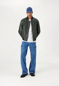 Donkergroene leren bomberjack, wit t-shirt, losse blauwe denim cargobroek met zijzakken, zwarte schoenen en een blauwe pet.