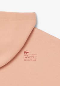 Hanorac roz piersici, cu o textură netedă, cusături bine încheiate și un logo Lacoste imprimat în roșu, alături de coordonatele locației.