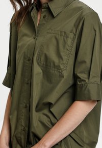 Camicia a maniche corte verde oliva con bottoni, dotata di taschino sul petto, realizzata in tessuto leggero e morbido, con colletto classico e vestibilità rilassata.