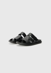 Black slide sandals made of textured material, featuring dual wide straps and an open toe design. Flat sole with subtle branding accents.