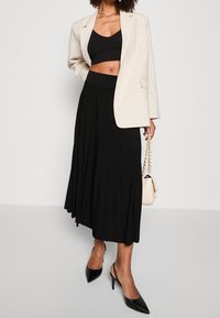 Blazer di lino beige su un top cropped nero, abbinato a una lunga gonna nera plissé e décolleté slingback nere con punta a stiletto. Borsa color crema con catena.