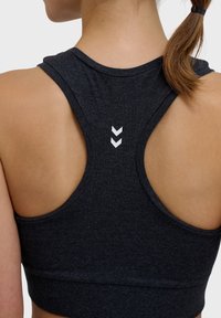 Mörkgrå atletisk crop top med racerback-design. Har en vit chevron-logotyp centrerad på ryggen. Slät, elastisk tygtextur.