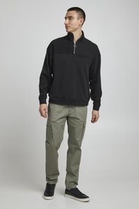 Svart quarter-zip hettegenser med teksturert øvre panel, paret med olivenfargede cargo-bukser med sidelommer og svarte sneakers.