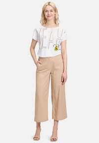 Witte crop top met "YEAH!" graphic en cartoonfiguur, gecombineerd met beige wijde broek en nude hakken. Gladde, lichte stof.