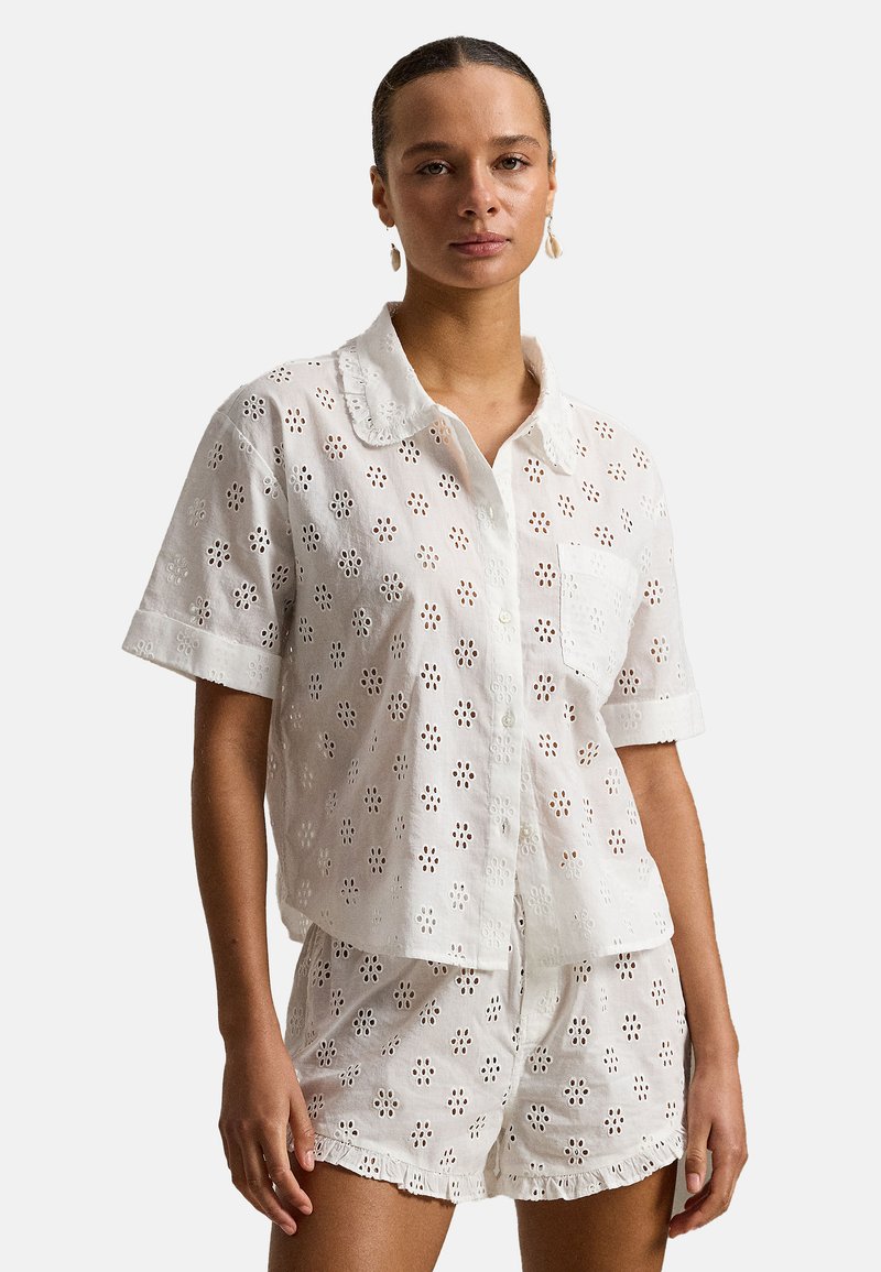 Vrouw draagt een wit shirt met knopen en bijpassende korte broek met bloemenmotief en opengewerkt patroon, staand tegen een effen achtergrond.