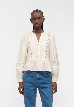 Femme aux cheveux tressés porte un chemisier crème orné de dentelle avec des boutons et des manches bouffantes, associé à un jean bleu, debout devant un fond blanc.