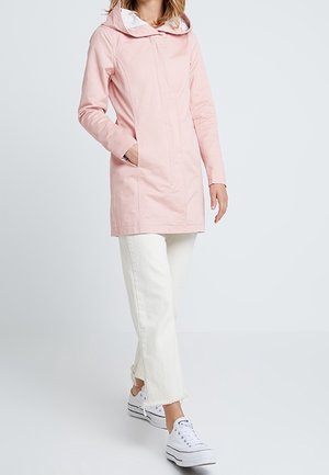 Personne portant un manteau à capuche rose clair, un pantalon blanc à ourlet effiloché et des baskets compensées blanches, marchant sur un fond clair uni.
