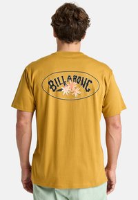 Gul bomulds T-shirt med en sort oval med ordet "BILLABONG" og hvide blomster grafikker på ryggen. Korte ærmer, afslappet pasform.