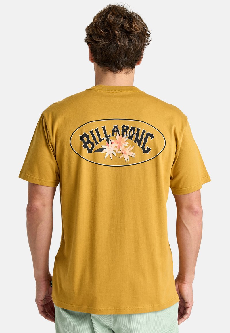Gul bomullströja med en svart oval med ordet "BILLABONG" och vita blomgrafik på baksidan. Korta ärmar, avslappnad passform.