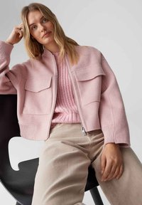 Veste courte rose en tissu doux avec deux poches poitrine, poignets et ourlet côtelés, portée sur un pull tricoté rose clair et un pantalon beige.