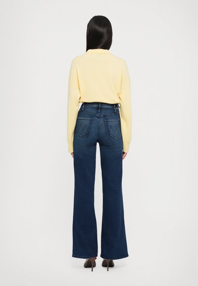 Jeans de talle alto de color azul oscuro con pierna acampanada, combinados con un suéter amarillo claro. Textura de denim suave y bolsillos traseros visibles.