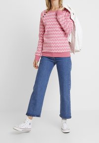 Pullover rosa con motivo a zigzag, caratterizzato da una scollatura tonda e bottoni su una spalla, abbinato a jeans di denim blu a gamba larga e sneakers bianche.