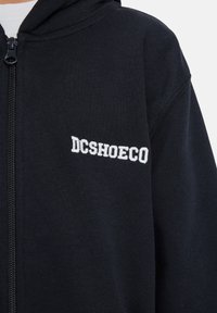 Čierna mikina na zips s bielym vyšitým nápisom "DCSHOECO" na ľavej hrudi, disponujúca textúrovanou tkaninou a viditeľným ťahákom na zips.