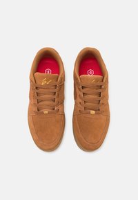 Paire de baskets à lacets en daim marron avec semelles intérieures rouges et coutures jaunes, vues de dessus sur fond blanc.