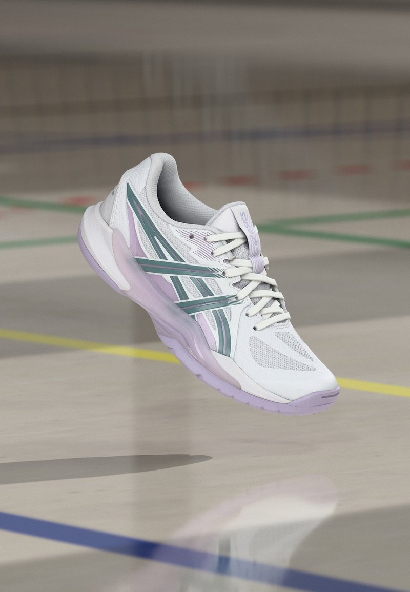 Chaussure de sport blanche et violette avec des bandes vertes en plein air au-dessus du sol d'un terrain de sport couvert avec des lignes colorées.