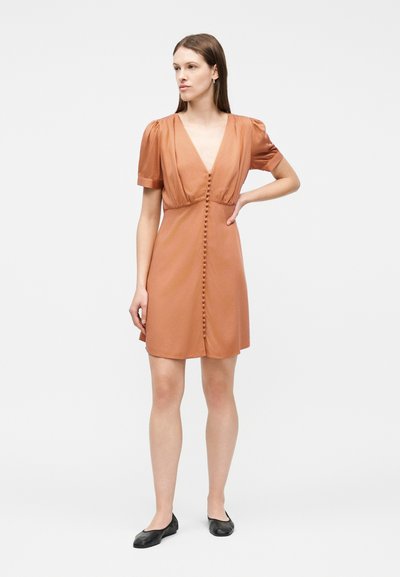 Γυναίκα που στέκεται με ένα χέρι στη μέση, φορώντας φορέμα κοντομάνικη, σε χρώμα σκουριάς με V-λαιμόκοψη και κουμπιά καλυμμένα με ύφασμα κατά μήκος μπροστά, καθώς και μαύρα φλατ παπούτσια.