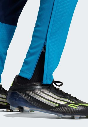 Sinised sportlikud püksid, millel on pahkluu piirkonnas luku detail, kombineeritud mustade ja hõbedaste jalgpallikingadega, millel on rohelised aktsendid ja tekstuuriga pinnad.