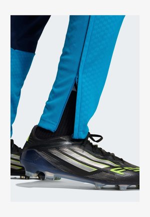 Blue athletic pants with a zipper detail at the ankle, paired with black and silver soccer cleats featuring green accents and textured surfaces.