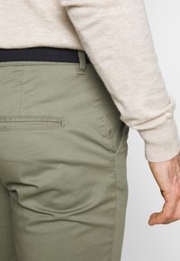 Kaki gröna byxor visade från baksidan, med en platt ficka och slät yta. Modellens ljusbeige tröja syns på överarmen.