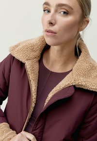 Burgundská bunda s béžovým umělým ovčím límcem; látka má hladkou texturu a je vybavena zipem vpředu a prošívanou podšívkou.