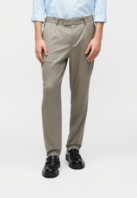 Άνδρας που φοράει ανοιχτό μπλε πουκάμισο, μπεζ ραμμένο παντελόνι και μαύρα δερμάτινα loafers, στέκεται μπροστά σε λευκό φόντο.