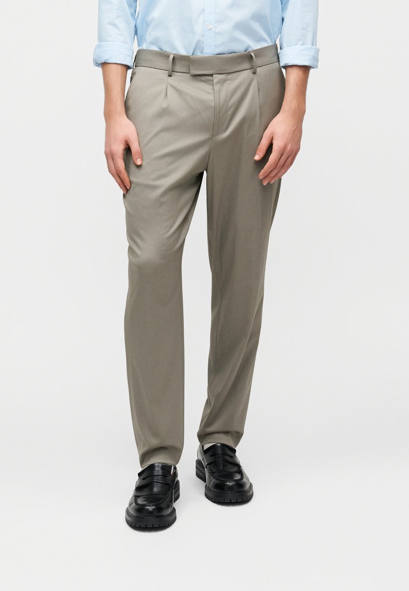 Άνδρας που φοράει ανοιχτό μπλε πουκάμισο, μπεζ ραμμένο παντελόνι και μαύρα δερμάτινα loafers, στέκεται μπροστά σε λευκό φόντο.