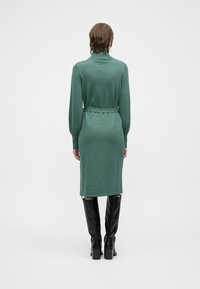 Strikket kjole i dempet grønn med lange ermer, høy krage, beltestropp i livet og rette linjer, parret med knelange svarte støvler.