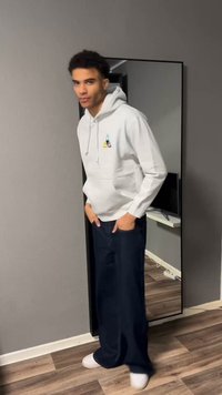 Giovane uomo con felpa bianca con cappuccio e jeans larghi scuri sta con le mani nelle tasche davanti a uno specchio a figura intera in una stanza.