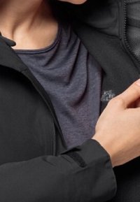 Svart zip-up-jacka med texturerat innerfoder, bärs över en mörkblå T-shirt. Närbild på ärm och halsområde som visar tyg och passformsdetaljer.