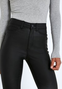 Svarta högmidjade jeans med en slät, glänsande yta, med standardfickor och knappstängning. Kombinerat med en ljusgrå, ribbad långärmad tröja.