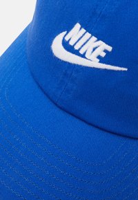 Mėlyna medvilninė beisbolo kepurė su struktūruotu karūna ir baltu siuvinėtu Nike logotipu priekyje, su tekstūruotu audiniu ir siuvimo detalėmis.