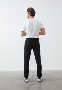 Joven con cabello de longitud media, vistiendo una camiseta blanca, jeans negros y zapatillas blancas, de pie frente a una pared blanca en interiores.