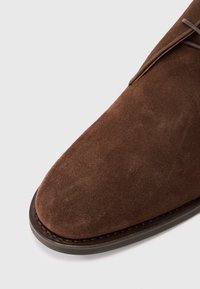 Primo piano della punta di una scarpa in camoscio marrone con suola cucita di un marrone scuro e lacci abbinati, su uno sfondo leggero e neutro.