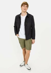 Sort zip-up jakke med to brystlommer og et logo-applikation, parret med olivengrønne cargo-shorts og hvide sneakers. En ensfarvet hvid t-shirt under.