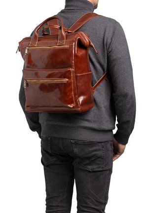 Zaino in pelle marrone con superficie texturizzata, diverse tasche con zip e doppi manici, indossato su un maglione grigio.