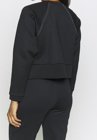Donna che indossa un top nero a maniche lunghe con dettagli di cucitura bianca e pantaloni neri aderenti, vista da dietro.