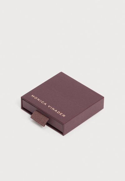 Μικρό τετράγωνο σκούρο καφέ κουτί κοσμημάτων με ταινία έλξης, με την επιγραφή "Monica Vinader" σε χρυσά γράμματα στο καπάκι, σε απλό ανοιχτό φόντο.