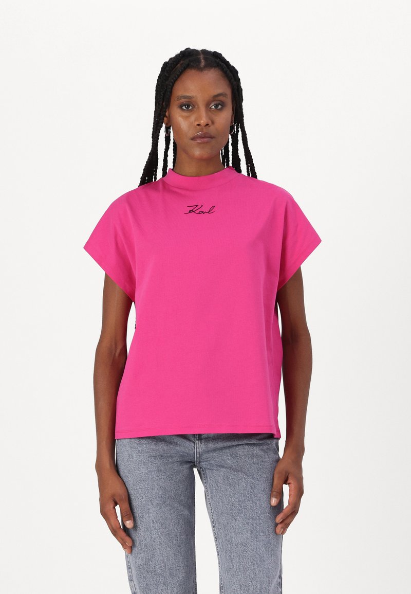 Rosa bomull t-skjorte med høy hals og korte ermer. Har en svart brodert logo på brystet. Kombinert med lyseblå jeans.