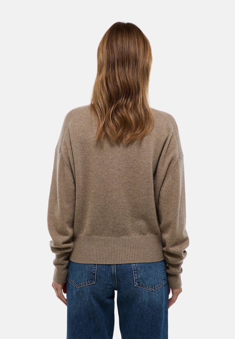 Personne avec des cheveux ondulés de longueur épaule, portant un pull beige et un jean bleu, se tenant de dos face à l'appareil photo.