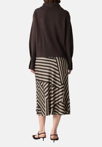 Brauner Strickpullover mit hohem Kragen, kombiniert mit einem knielangen Kleid, das auffällige, kantige beige und dunkelbraune Streifen aufweist.