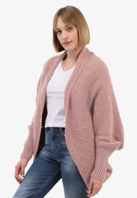 Frau, die eine strukturierte pinke Strickjacke über einem weißen T-Shirt und blauen Jeans trägt und vor einem einfarbigen hellen Hintergrund posiert.