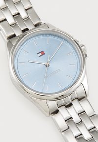 Silver stainless steel watch with a round case. Light blue dial, silver hour markers, and a small logo. Metal link bracelet with polished finish.