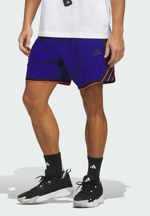 Mężczyzna w granatowo-czarnych sportowych szortach z pomarańczowym wykończeniem, czarnych skarpetkach Adidas oraz czarno-białych sportowych butach, stoi z lekko rozstawionymi nogami.