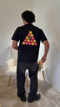 Άτομο με σγουρά μαλλιά στέκεται με την πλάτη, φοράει μαύρο πουκάμισο και παντελόνι. Στη πίσω πλευρά του πουκαμίσου υπάρχει πολύχρωμο κυκλικό σχέδιο με γράμματα.