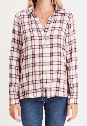 Femme portant une chemise à carreaux crème, bordeaux et bleu marine à manches longues et un jean bleu, debout devant un fond blanc.
