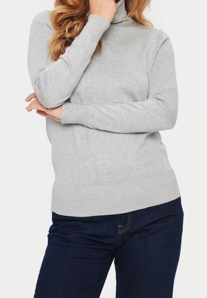Femme portant un pull à col roulé gris clair et un jean bleu foncé, une main touchant son menton et l'autre croisée sur sa taille.