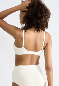 Reggiseno bianco realizzato in materiale morbido, con spalline regolabili e chiusura con gancio e occhiello. Mutandine a vita alta abbinate visibili sotto.