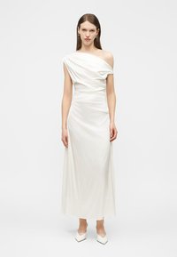 Nő, fehér egyrészes szatén ruhában, testhezálló derékkal és fehér hegyes orrú magassarkú cipőben, egy egyszerű háttér előtt áll.