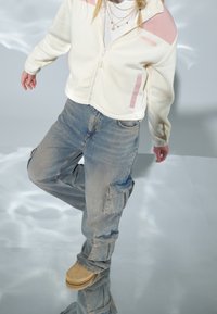 Henkilö, jolla on valkoinen ja vaaleanpunainen fleecevetoketjulla, kerroksittain kaulakoruja, löysät siniset cargo-farkut ja ruskeat saappaat, seisoo heijastavalla pinnalla.