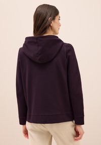 Felpa viola con una texture morbida, dotata di cappuccio e polsini a costine, abbinata a pantaloni di colore chiaro.